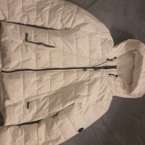 Vit quiltad pufferjacka med huva - Säljer en snygg vit pufferjacka med quiltat mönster och svart dragkedja. Jackan har huva med justerbar snörning och praktiska fickor med svarta detaljer. Perfekt för kalla dagar och riktigt skön att bära.