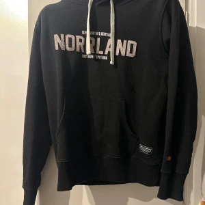 Svart hoodie från SQRTN Company XS - Svart hoodie från SQRTN Company med vit broderad text 'NORRLAND' på bröstet. Klassisk känguruficka framtill, vita snören i huvan och ribbade muddar. Perfekt för dig som gillar streetwear och vill ha en stilren look.