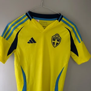 Svenska landslagets fotbollströja Adidas - Gul fotbollströja från Adidas med blå detaljer och Sveriges landslagsemblem på bröstet. Klassiska tre ränder på axlarna, rund hals med blå kant och texten 'Sverige' på ryggen. Tillverkad i lätt och ventilerande material, perfekt för match eller träning.