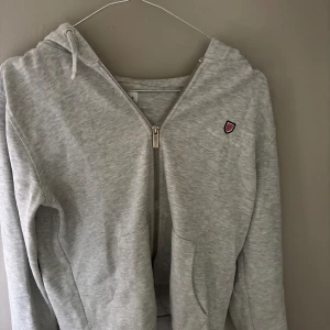 Ljusgrå hoodie från Bondelid  - Ljusgrå hoodie från Bande Girl med dragkedja framtill, broderad logga på bröstet och två fickor. Tröjan har huva med snörning och är tillverkad i mjuk bomullsblandning. Perfekt för chill dagar eller när du vill vara extra bekväm. Ärmarna är lite trasiga 