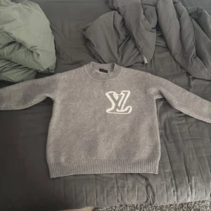 Grå stickad tröja Louis Vuitton - Snygg grå stickad tröja från Louis Vuitton med stort vitt LV-logo både fram och bak. Tröjan har rund hals, ribbade muddar och är långärmad. Perfekt för dig som vill ha en clean och exklusiv look.