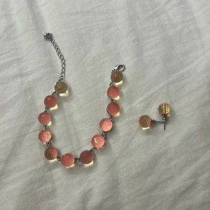 Armband och örhängen i rosa och gult - Set med armband och matchande örhängen i silverfärgad metall. Båda har runda, glittriga plattor i rosa och gult med en skimrande, nästan holografisk yta. Armbandet har justerbar kedja och örhängena är små och diskreta med samma färgade plattor.