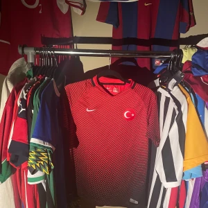Turkiet fotbollströja Nike 158-170, äkta - Säljer en officiell Turkiet fotbollströja från Nike i rött med svart rutmönster. Tröjan har korta ärmar, Dri-FIT-material som andas och Turkiets emblem på bröstet. Perfekt för dig som vill sticka ut på planen eller visa ditt stöd för landslaget. Äkta