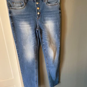 Blå raka/skinny jeans med knappar - Snygga ljusblå raka/skinny jeans med fyra silverfärgade knappar framtill och slitna detaljer på både fram- och baksida. Klassisk femficksmodell med bälteshällor och lätt tvättad look. Perfekta för en avslappnad och trendig stil.