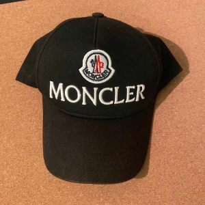Svart keps från Moncler - Svart keps från Moncler med stor vit broderad logotyp och text framtill. Klassisk böjd skärm och justerbar passform. Tillverkad i bomull för en bekväm känsla och stilren look.