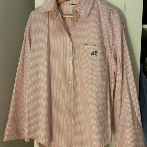 Randig rosa skjorta från Gina Tricot - Säljer en oversized rosa skjorta från veromoda med vita ränder. Skjortan har klassisk krage, bröstficka med broderad logga och knäppning framtill. Långärmad och tillverkad i mjuk bomull. Perfekt för en chill och trendig look.