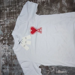 Vit t-shirt från AMI Paris med rött hjärta - Vit t-shirt från AMI Paris med klassisk passform och rund hals. På bröstet sitter ett stort rött hjärta med bokstaven A broderat. Tillverkad i mjuk bomull och har korta ärmar. Perfekt statement-plagg för dig som gillar stilrena men unika detaljer.