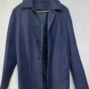 Mörkblå Kappa från ASOS - Säljer en stilren mörkblå kappa från ASOS med klassisk krage och knäppning framtill. Jackan har raka fickor med diskreta detaljer och är helfodrad för extra komfort. Perfekt för dig som gillar en clean och tidlös look.