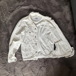 Vit jeansjacka från Zara, storlek S - Säljer en vit jeansjacka från Zara i storlek S. Jackan har slitna detaljer vid krage och axlar, klassisk knappstängning framtill och två bröstfickor med lock. Perfekt för dig som gillar en avslappnad och trendig look.