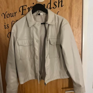 Beige skinnjacka med fickor - Snygg beige skinnjacka med klassisk krage och två stora bröstfickor med lock och tryckknappar. Jackan har dragkedja framtill och raka ärmar med knappdetalj vid ärmslut. 