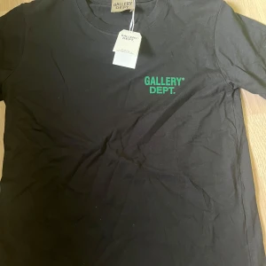 Svart t-shirt från Gallery Dept. - Svart t-shirt från Gallery Dept. med grönt tryck fram och stort tryck på ryggen med texten 'GALLERY DEPT. HOLLYWOOD, CA.'. Klassisk rund hals och korta ärmar. Skön bomullskvalitet och clean streetwear-vibe.