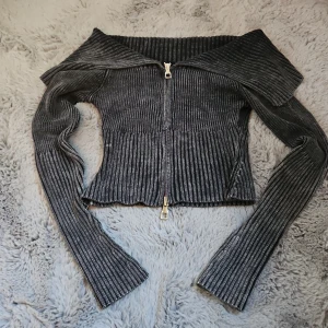 Offshoulder kofta - Snygg mörkgrå ribbad kofta med offshoulder-design och långärmade ärmar. Tröjan har en dragkedja framtill och är croppad i modellen, vilket ger en trendig look. Perfekt för dig som gillar stickade detaljer och vill ha något som sticker ut.