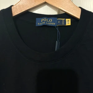 Svart t-shirt från Polo Ralph Lauren - Svart t-shirt från Polo Ralph Lauren med klassisk rund hals och korta ärmar. T-shirten har en röd broderad logga på bröstet och är tillverkad i mjuk bomull. Enkel och stilren modell som passar till det mesta.