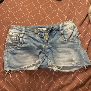 Blå jeansshorts från Oxygene - Snygga ljusblå jeansshorts från Oxygene med råa, fransiga kanter och slitna detaljer. Klassisk femficksmodell med knapp och dragkedja framtill. Perfekta för en avslappnad och cool stil under varma dagar.