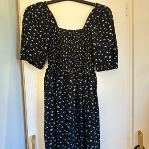 Svart blommig kortklänning Stories - Supersöt svart kortklänning från Stories med vita små blommor. Klänningen har puffiga korta ärmar, smockad överdel och fyrkantig urringning. Materialet är mjukt och lätt, perfekt för varma dagar. Passformen är normal och klänningen har en flowy siluett.