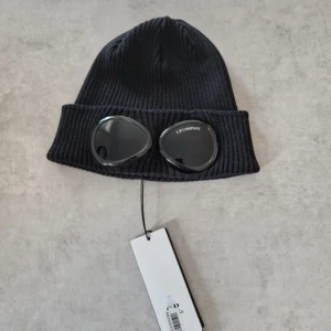 Svart Goggle Beanie från C.P. Company - Svart ribbstickad mössa från C.P. Company med ikoniska goggle-detaljer framtill. Tillverkad i mjuk bomull och har en uppvikt kant med två svarta glasögonlinser, varav ena är märkt med C.P. Company.                                                     • Helt ny                                                                   •Storlek Onesize.                                                 • Köpt från NK Nypris 1599kr