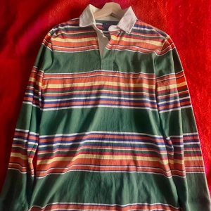 Randig pikétröja från Polo Ralph Lauren - Säljer en klassisk randig pikétröja från Polo Ralph Lauren i grönt, orange, blått, vitt, rött och gult. Skjortan har vit krage, långa ärmar och är tillverkad av bomull. Perfekt för dig som gillar preppy stil och vill sticka ut med färg.