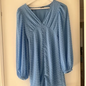 Blå kortklänning från Vero Moda - Supersöt blå klänning från Vero Moda med prickigt mönster och v-ringad hals. Klänningen har långa puffärmar med resår vid ärmslut och dekorativa knappar framtill. 💞