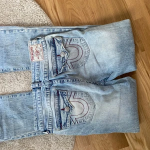 True Religion ljusa bootcut jeans - Säljer ett par ljusblå jeans från True Religion med bootcut passform. Jeansen har slitna detaljer på benen, klassiska bakfickor med röd kontrastsöm och råa kanter vid bensluten. Ikonisk True Religion patch baktill och knappar på fickorna. Perfekta för en avslappnad streetstil. Jag är 164 och dem är för långa 