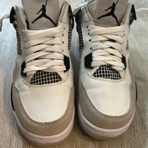 Nike Air Jordan 4 vit/svart/grå - Säljer ett par Nike Air Jordan 4 sneakers i vitt, svart och grått. Skorna har klassisk meshpanel, snörning och detaljer i mocka och skinn. Ikonisk Jumpman-logga på plösen och hälen. Perfekta för dig som vill sticka ut med en tidlös och sportig look.