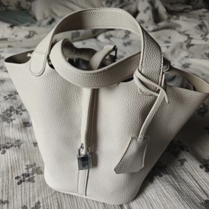 Vit bucket handväska i vegansk skinn - Fin handväska, vegan, fin material. Får plats med mycket, bärs i handen eller vid armbågen. Ingår nyckel och lås. Använd ett par gånger. Medföljer ett band man kan knäppa så att man kan bära den vid axeln. Inte exakt samma vita och lite mer guldig metalldelar men det fungerar. .