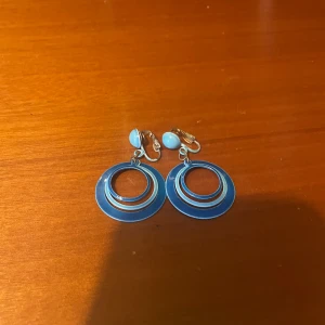 Blå retro örhängen med cirklar - Säljer ett par coola blå retro örhängen med cirkelformad design och clip-on fäste. Örhängena har flera ringar i olika storlekar som ger en snygg 70-talsvibe. Perfekt för dig som vill sticka ut med färg och form!