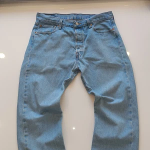 Levi's 501 ljusblå jeans W34 L34 - Klassiska Levi's 501 jeans i ljusblå denim. Rak passform med fem fickor, knappgylf och ikonisk läderpatch bak i midjan. Tillverkade i 100% bomull för en autentisk jeanskänsla. Perfekta för dig som gillar tidlös stil och vill ha ett par riktigt schyssta jeans.