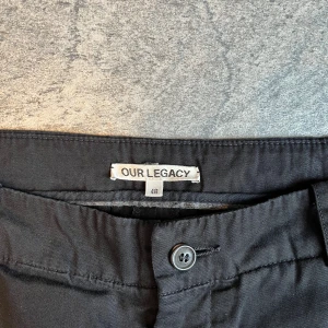 Svarta kostymbyxor från Our Legacy - Svarta kostymbyxor från Our Legacy i rak modell med klassisk knappgylf och bälteshällor. Byxorna har två bakfickor med knappar och en justerbar spänne bak. Tillverkade i ett mjukt bomullstyg som känns skönt mot huden.