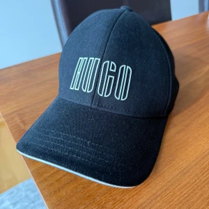 Svart keps från Hugo Boss - Svart keps från Hugo Boss med vit logotyp framtill i modern, grafisk stil. Justerbar rem baktill med metallspänne. Klassisk böjd skärm och ventilerande hål. Tillverkad i bomull för en bekväm och snygg look.