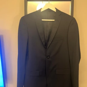 Svart kavaj Dressmann slim fit 42R - Svart kavaj från Dressmann i slim fit-modell, storlek 42 Regular. Klassisk design med smal passform, enkel knappstängning och två framfickor med lock. Perfekt för dig som gillar stilrena och moderna kostymer.