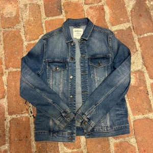 Blå jeansjacka från Dobber - Klassisk blå jeansjacka från Dobber i storlek L. Jackan har två bröstfickor med lock och knappar, samt snygga sömdetaljer och knäppning framtill. Perfekt för dig som gillar en tidlös och avslappnad stil.