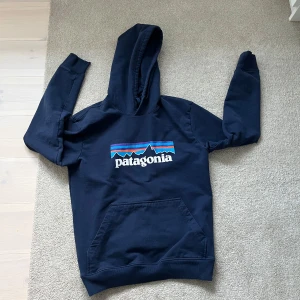 Mörkblå Patagonia hoodie med tryck - Säljer en mörkblå hoodie från Patagonia med klassiskt logotryck i vitt, blått, lila och orange på bröstet. Tröjan har huva, lång ärm och en stor magficka framtill. Perfekt för dig som gillar streetwear och outdoor vibes.