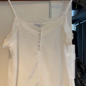 Vit spetsdetaljerad linnetopp Chiquelle - Supersöt vit linnetopp från Chiquelle med smala axelband, spetskant upptill och dekorativa knappar framtill. Toppen har en feminin v-ringning och är tillverkad i ett lätt material som passar perfekt till varma dagar. Spetsen ger en romantisk touch och toppen är ärmlös.