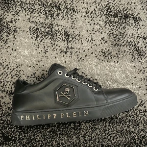 Svarta sneakers från Philipp Plein - Snygga svarta sneakers från Philipp Plein i skinn med cool döskalle-logga på sidan och metallplatta på plösen. Skon har svarta snören med logga och PHILIPP PLEIN i silver på sulan. Perfekt för dig som gillar statement-detaljer och exklusiv streetstyle.