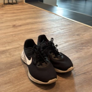 Svarta Nike sneakers strl 42/41  - Säljer ett par svarta Nike sneakers med vit sula och vit swoosh-logga på sidan. Skorna har snörning och är tillverkade i mesh och syntetmaterial, vilket gör dem lätta och sköna för vardagsbruk eller träning. Klassisk och stilren design som passar till det mesta. De är ganska små i storleken så skulle säga att de mer är storlek 41 än 42. 
