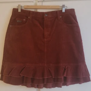 Roströd manchesterkjol Odd Molly - Supersnygg roströd manchesterkjol från Odd Molly med volangkant nertill och klassiska jeansfickor fram och bak. Kjolen har fina metallknappar och detaljer, samt bälteshällor. Perfekt för dig som gillar retrovibbar och vill sticka ut lite extra.