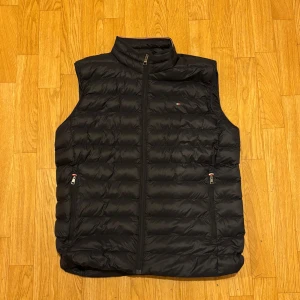 Svart dunväst från Tommy Hilfiger - Svart dunväst från Tommy Hilfiger med quiltad design och dragkedja framtill. Västen har två fickor med dragkedja och liten logga på bröstet. Hög krage och lätt att packa ner. Perfekt för kyliga dagar när du vill hålla stilen.