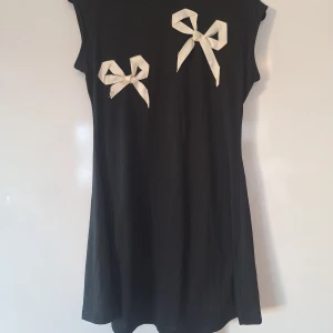 Svart kort klänning med rosetter från Cider S/M - Säljer en svart kort klänning med två vita rosetter framtill. Klänningen är ärmlös och har en enkel, rak passform som ger en chill vibe. Perfekt för dig som gillar stilrena detaljer och vill sticka ut med något unikt.