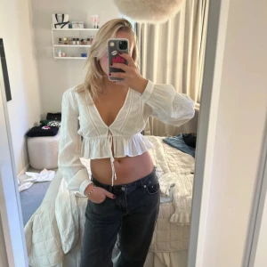 Vit croppad blus med knyt - Säljer en superfin vit blus med v-ringning och knyt framtill. Blusen är croppad med volangkant och har långa, lätt puffiga ärmar i tunt bomullstyg med broderade detaljer. Perfekt för en fräsch och somrig look! Passar även S!