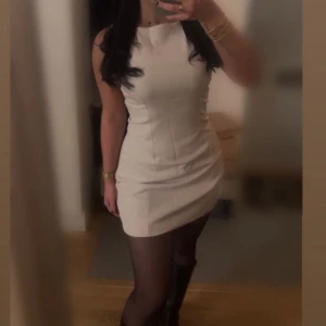 Beige kortklänning med hög hals - Säljer en stilren beige kortklänning med hög hals och figurnära passform. Klänningen är ärmlös och har en enkel, clean design som ger en snygg siluett. 