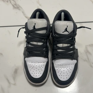 Nike Air Jordan 1 Low svart/vit - Säljer ett par Nike Air Jordan 1 Low i svart och vitt skinn med klassisk Jumpman-logga på plösen och Air Jordan Wings på hälen. Skorna har svarta snören, perforerad tåbox och en svart sula. Perfekt för dig som gillar streetwear och vill ha en ikonisk sneaker i garderoben.