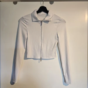 Vit ribbad croppad kofta med dragkedja - Säljer en vit croppad kofta med ribbad struktur och lång ärm. Tröjan har en klassisk krage och hel dragkedja framtill. Perfekt att styla med jeans eller kjol för en clean look.
