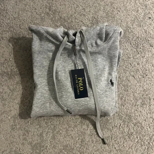 Grå hoodie från Polo Ralph Lauren - Snygg grå hoodie från Polo Ralph Lauren med klassisk broderad logga på bröstet. Tröjan har huva med snörning, känguruficka och ribbade muddar. Perfekt för chill dagar och enkel att matcha med jeans eller joggers.