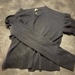 Svart kofta med volangärmar - Mysig svart kofta med volangdetaljer på ärmarna. Klassisk rund halsringning och ribbade muddar vid ärmslut och nederkant. 