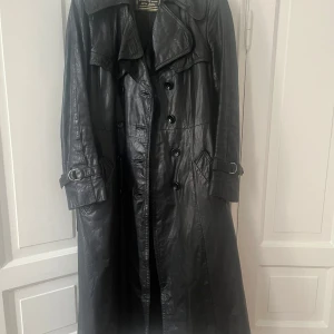 Svart lång skinn trenchcoat - Säljer en svart lång trenchcoat i äkta skinn från HM. Jackan har dubbelknäppning, bred krage, bälte i midjan och spännen vid ärmsluten. Perfekt för dig som vill ha en edgy och tidlös look.