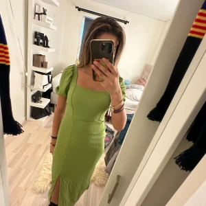 Grön långklänning med puffärm och snörning - Säljer en snygg grön långklänning med puffärmar, fyrkantig ringning och slits framtill. Klänningen har öppen rygg med snörning och är gjord i ett strukturerat, stretchigt material. Perfekt för dig som vill sticka ut med en cool och trendig look!