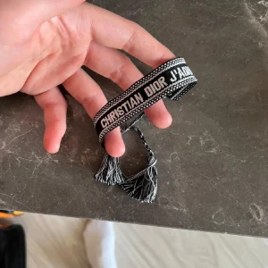 Christian Dior svart/vitt armband - Snyggt armband från Christian Dior i svart och vitt med texten 'CHRISTIAN DIOR J'ADIOR'. Armbandet har tofsar i ändarna och är vävt i tyg med coolt mönster. Perfekt accessoar för att lyfta din outfit.