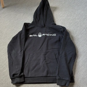 Sail Racing hoodie Junior med tryck  - Svart hoodie från Sail Racing (junior)  med vit logga framtill. Klassisk känguruficka, huva och ribbade muddar. Mjuk insida och långärmad modell, perfekt för chill dagar eller när du vill ha en clean streetstyle.