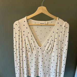 Vit prickig v-ringad topp Kappahl - Vit ribbad topp från Kappahl med svarta prickar och djup v-ringning. Långärmad modell med lätt rynkade detaljer vid bysten för en snygg passform. Perfekt för dig som gillar en klassisk men trendig look.