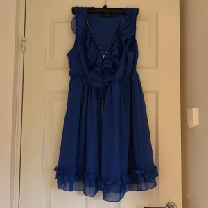 Blå volangklänning med dragkedja - Supersöt blå kortklänning från Vila med volanger längs v-ringningen och nederkanten. Klänningen är ärmlös och har en synlig dragkedja framtill. Tillverkad i ett lätt och luftigt polyestertyg som ger en flowy känsla.